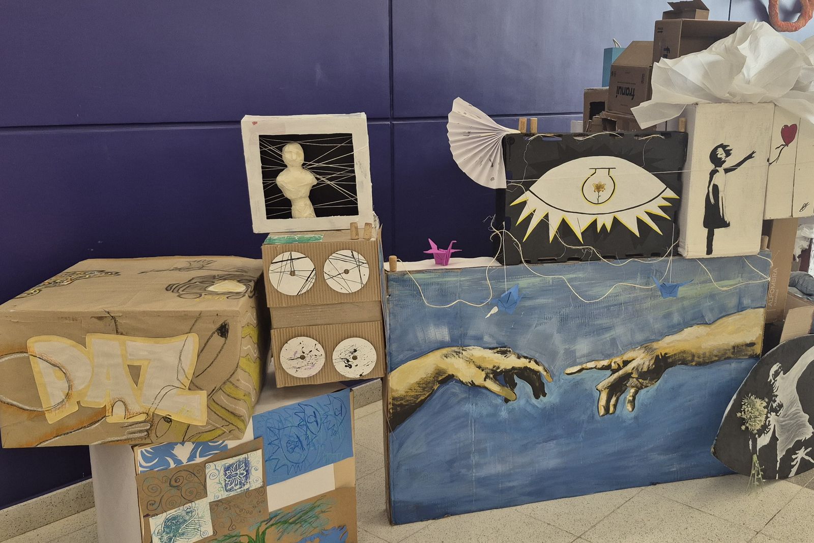 Obras de la Expo Arte 2026: una instalación de cartón con dos manos pintadas sobre fondo azul, junto a otras piezas de estudiantes